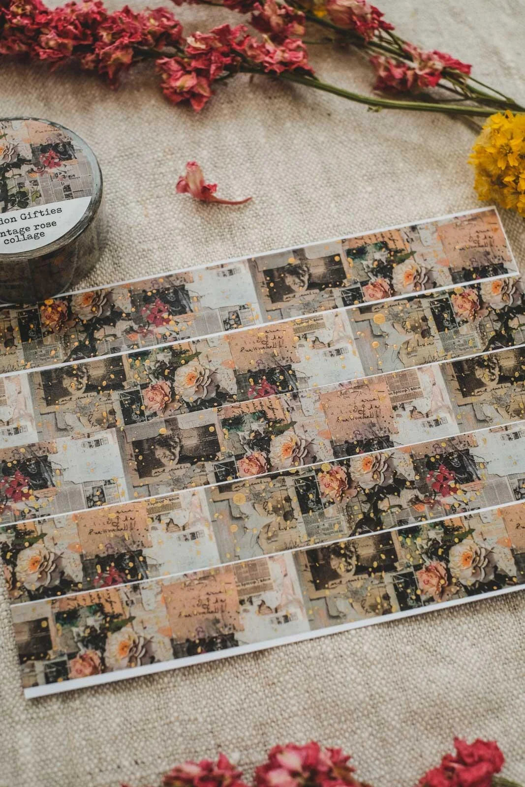 A styled flat-lay of decorative washi tape strips featuring a vintage collage pattern with floral illustrations, handwritten postcards, and muted cityscapes. The tape is laid out on a beige linen fabric alongside dried pink flowers, a small container of washi tape, and yellow flowers.