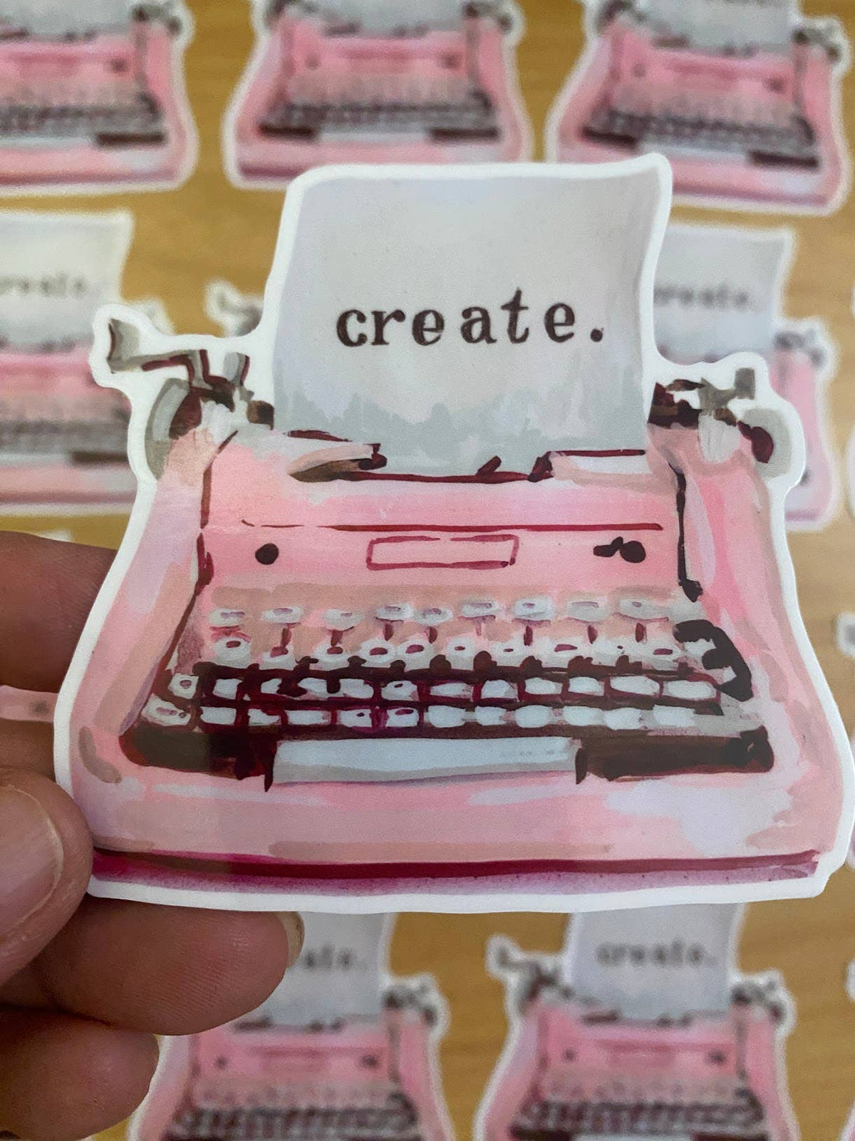A hand holds a die-cut sticker featuring a pink watercolor-style typewriter with the word 'create.' on the paper.