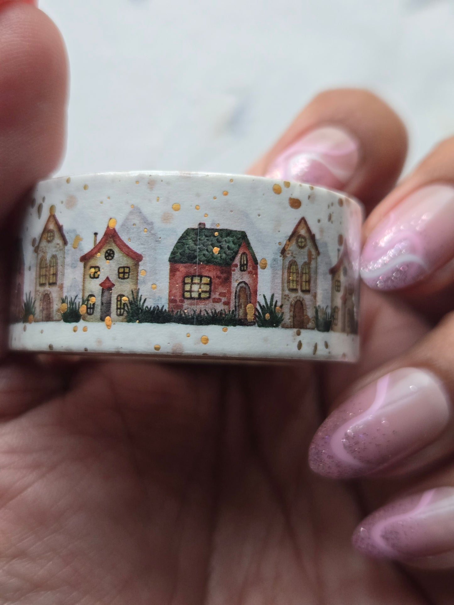 A close-up of a hand holding a roll of washi tape featuring a winter village scene with small houses, trees, and gold speckles on a white background.