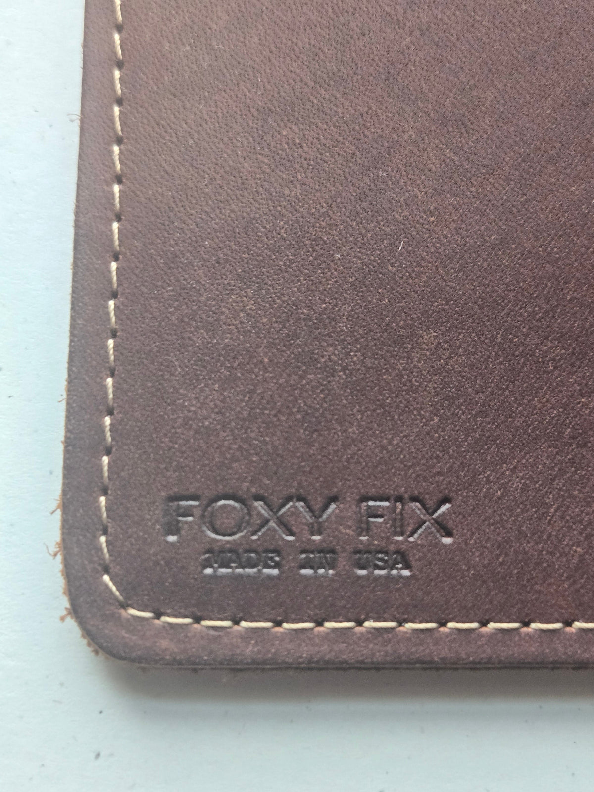 Close-up of a brown leather wallet corner with white stitching and an embossed 'FOXY FIX' logo on a light blue background.