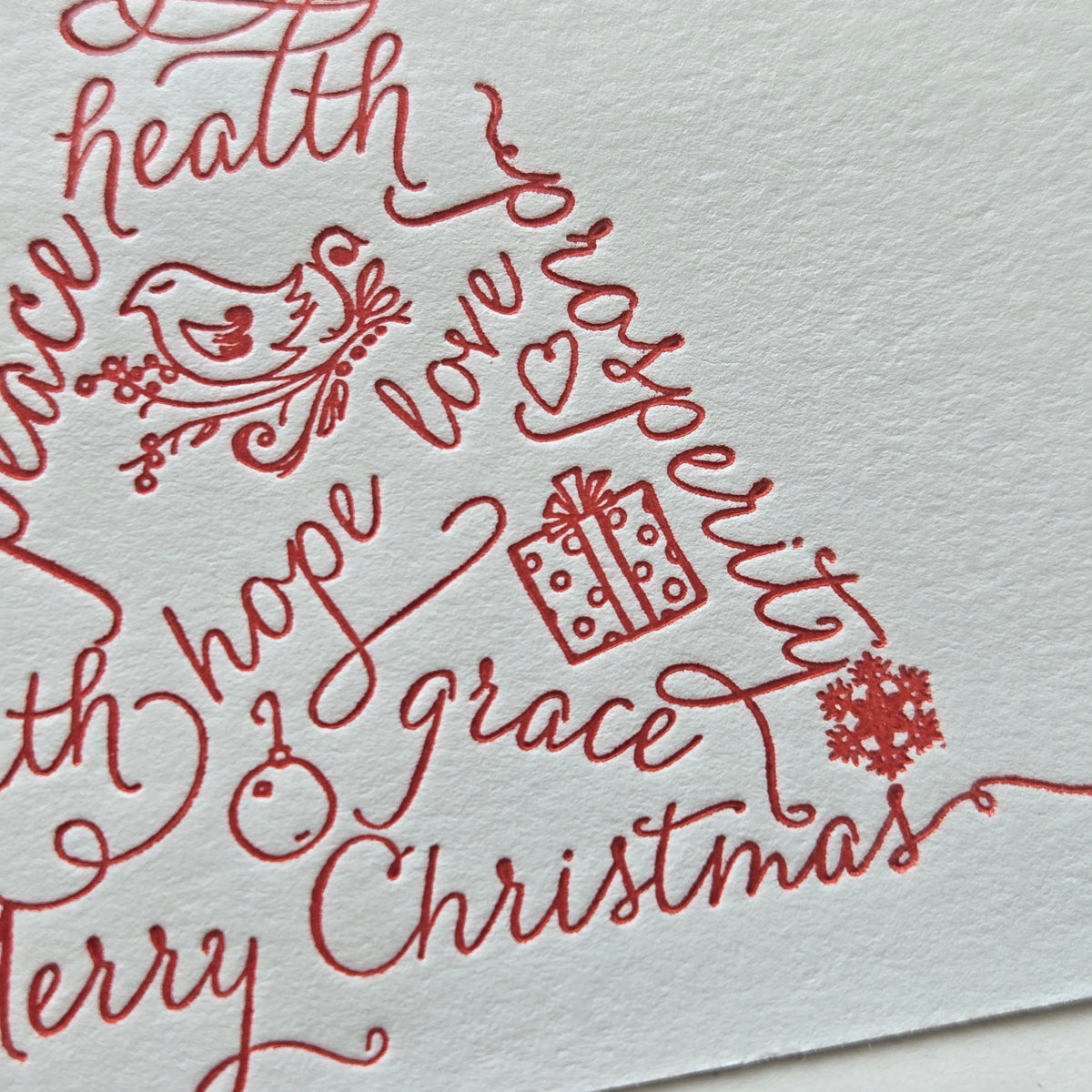A close-up of a white textured paper card featuring elegant red cursive holiday lettering that reads 'health prosperity hope grace Christmas' with decorative illustrations of a bird, gift, and snowflake.