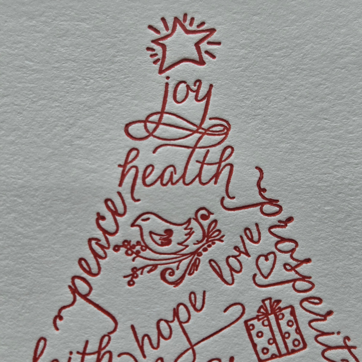 A close-up of a textured light gray paper with red hand-lettered script forming a Christmas tree shape, decorated with words like joy, health, peace, hope, love, and faith, plus a star, bird, and gift icon.