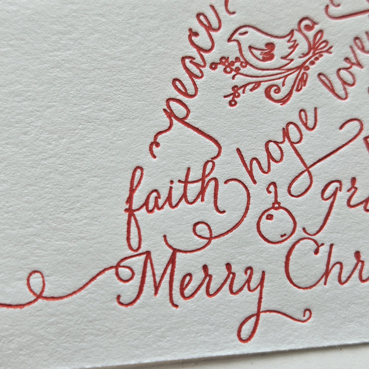 Close-up of red calligraphic holiday lettering on textured white paper, including the words 'faith', 'hope', 'gratitude', and 'Merry Christmas' with a small bird motif near the top.