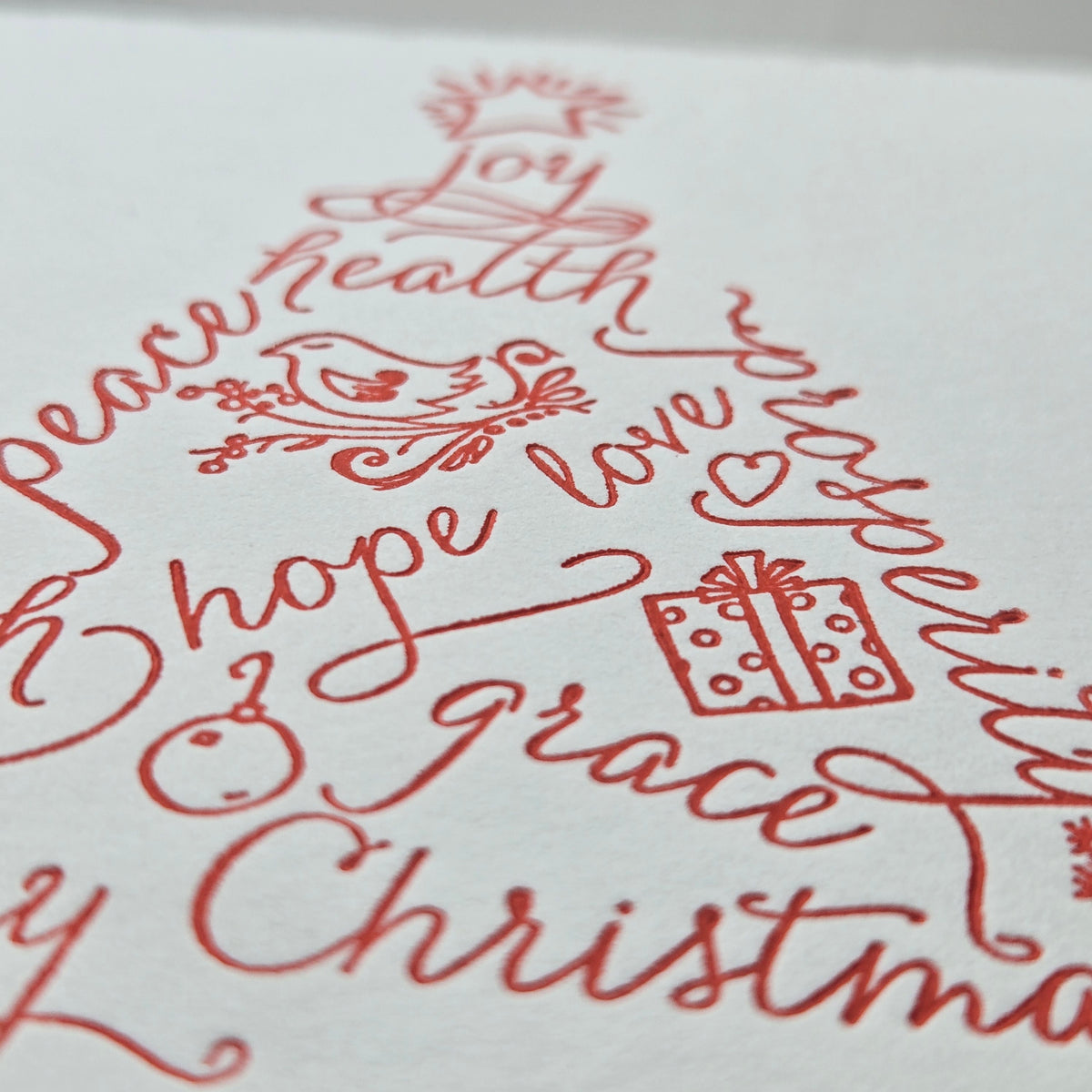 A close-up of red calligraphic Christmas lettering on white paper, forming festive words and motifs including 'Joy', 'health', 'prosperity', 'peace', 'hope', 'love', 'grace', and 'Christmas'.
