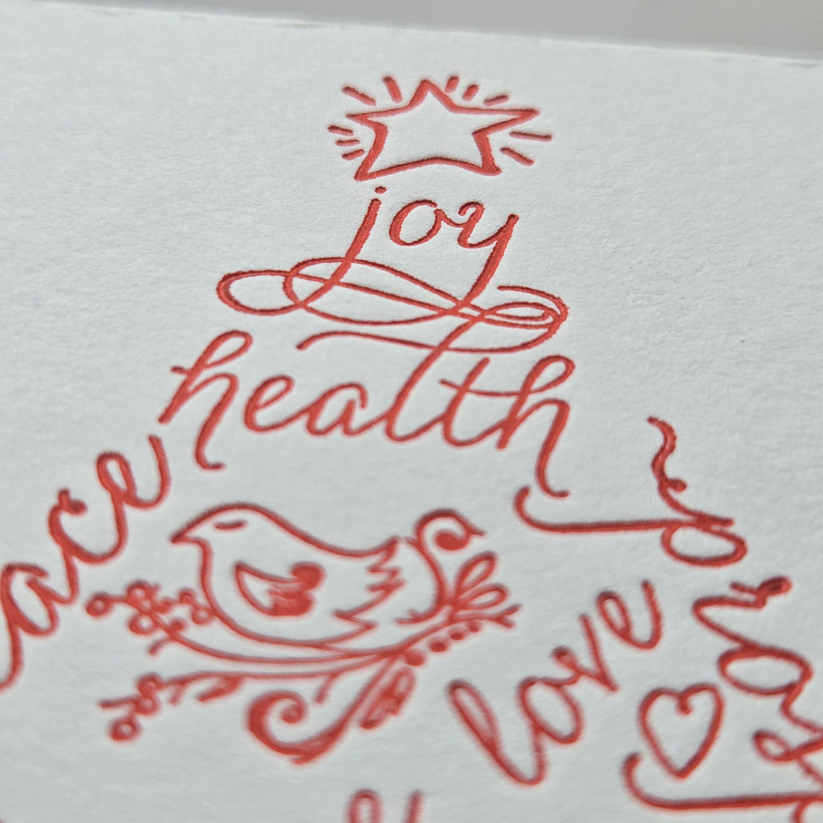 A close-up of a white textured paper card featuring elegant red cursive script that spells out 'health', 'love', and 'joy' in the shape of a Christmas tree, decorated with a red star and bird motif.