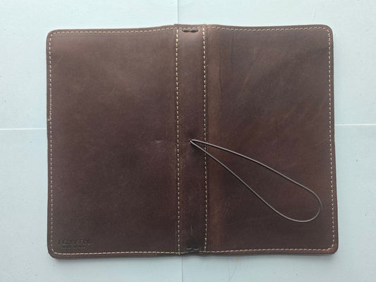 A brown leather card holder wallet with a loop strap, laid flat on a light gray tiled surface.