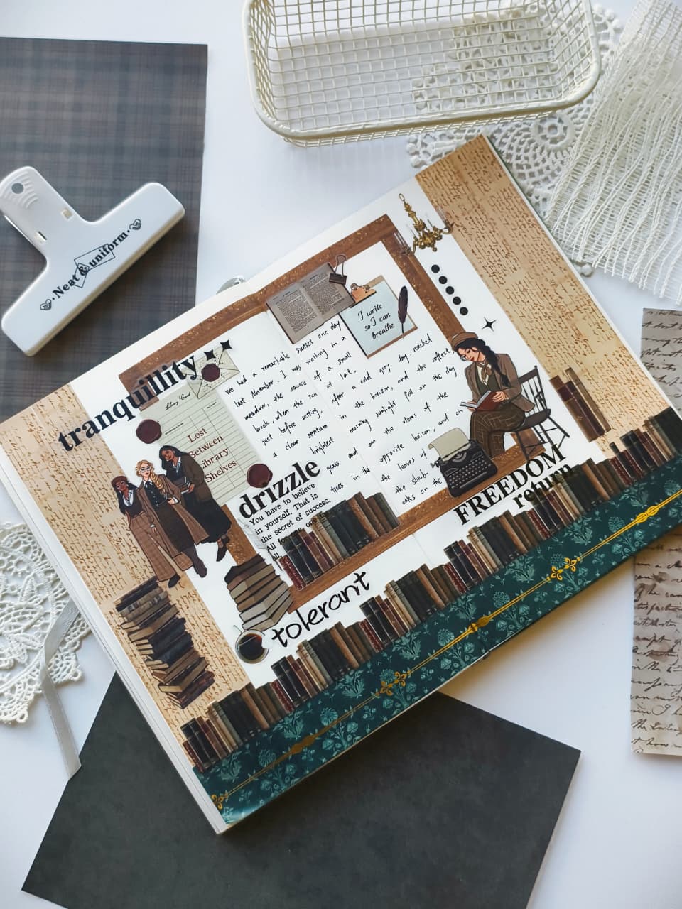 A decorative scrapbook journal or book cover laid flat on a white surface, surrounded by paper clips, lace, and patterned paper. The cover features a vintage bookish collage with illustrated figures, book spines, handwritten text, and words including 'tranquility,' 'drizzle,' 'tolerant,' and 'freedom.