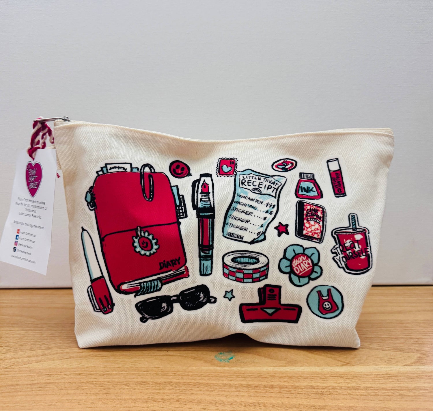 An off-white canvas zippered pouch featuring a colorful graphic print of various stationery and beauty items, including a red backpack, lipstick, sunglasses, and a receipt, resting on a wooden surface against a plain light gray background.