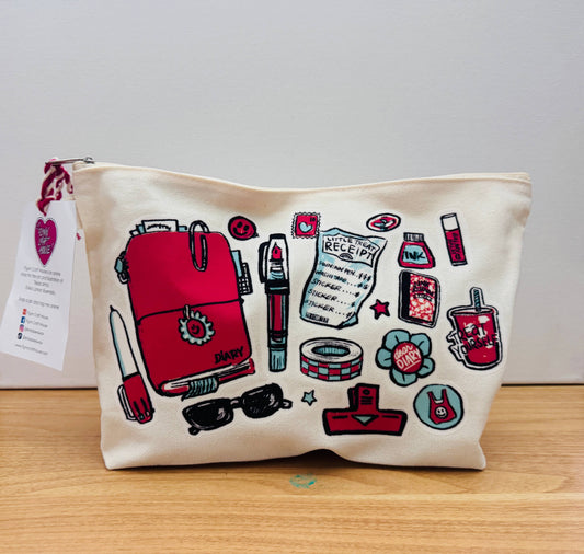 An off-white canvas zippered pouch featuring a colorful graphic print of various stationery and beauty items, including a red backpack, lipstick, sunglasses, and a receipt, resting on a wooden surface against a plain light gray background.