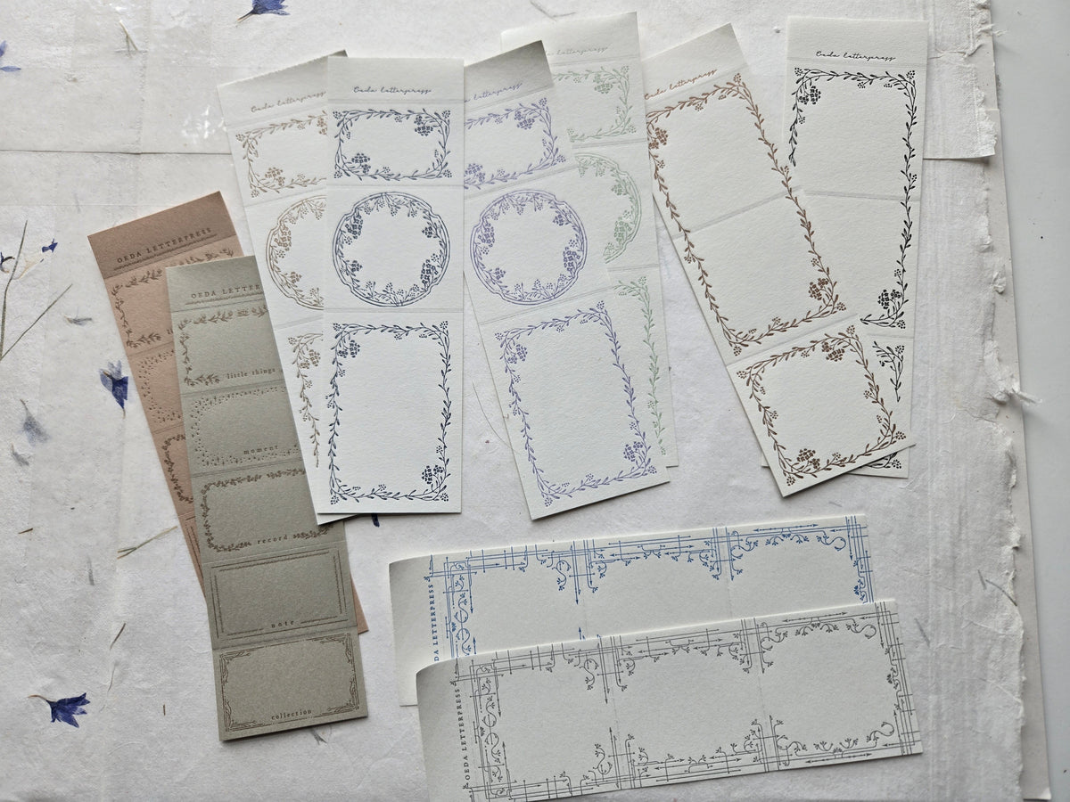 A flat lay of multiple sheets of decorative paper with ornate vintage-style border frames in muted blue, gray, beige, and mauve tones, arranged on a textured off-white fabric background.